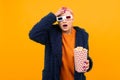 Extraordinary beautiful woman with short pink hair in dark coat and glasses with popcorn isolated on orange background