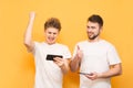 Extreme men emotionally play games on a smartphone, stand on a yellow background, wear white t-shirts. Two people are emotionally