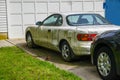 An extremely dirty white car parked in a driveway by a multi-panel garage door