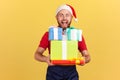 Extremely happy courier in blue uniform and red santa claus hat holding and showing christmas gifts looking at camera with toothy