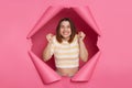 Extremely happy enjoyable brunette woman wearing striped top looking through ripped pink paper hole raised her arms clenched fists Royalty Free Stock Photo