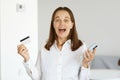 Extremely happy excited woman wearing white shirt standing indoor in light room with smart phone and credit card in hands, winning Royalty Free Stock Photo