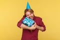 Extremely happy guy with funny party cone on head embracing gift box, shouting for joy excitement, indoor