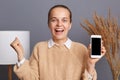 Extremely happy joyful woman wearing beige sweater posing in stylish room standing showing mobile phone with empty display, Royalty Free Stock Photo