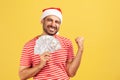 Extremely happy pleased bearded man in red t-shirt and santa claus hat showing yes gesture holding fan of dollars, perks and Royalty Free Stock Photo