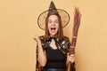 Extremely happy positive overjoyed young woman wizard wearing witch costume holding in hand broom  over beige background Royalty Free Stock Photo