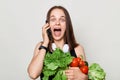 Extremely surprised overjoyed adult woman with brown hair standing isolated over white background holding vegetables talking on