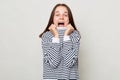 Extremely surprised positive overjoyed woman with brown hair wearing striped shirt standing isolated over gray background