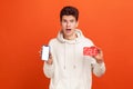 Extremely surprised teenager in casual hoodie widely open eyes and mouth holding sale card and smartphone in hands, shocked with