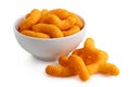 Extruded cheese puffs in a white ceramic bowl next to spilled cheese puffs isolated on white