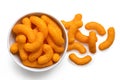 Extruded cheese puffs in a white ceramic bowl next to spilled cheese puffs isolated on white. Top view