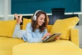 Exuberant girl uses tablet pc while laying on the comfortable yellow sofa and enjoying nice melodies with earphones.