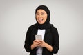 An exuberant young woman in a black hijab and blazer holds a smartphone with both hands Royalty Free Stock Photo