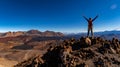 An exultant hiker stands with arms raised atop a rugged volcanic summit