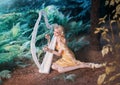 Fabulous forest elf sits under tree and plays on white harp, girl with long blond hair braided in long yellow dress Royalty Free Stock Photo