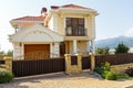 The facade of a beautiful white two-story cottage with a red roof. The cottage is located behind a wooden fence