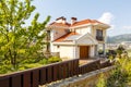 The facade of a beautiful white two-story cottage with a red roof. The cottage is located behind a wooden fence