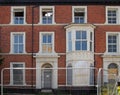 The facades of large abandoned derelict brick houses on a street with boarded up doors and windows behind a fence in southport