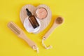 Face care set, cosmetic clay, wooden massage brush and amber dropper bottle, yellow background top view
