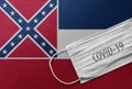 Face Medical Surgical White Mask with COVID-19 inscription lying on Mississippi State Flag. Coronavirus in Mississippi