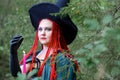 The face of the Sorceress with red hair in a hat and a black cloak in the woods in the hand with a candle.