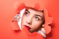 The face of a young beautiful girl with a bright make-up and with plump red lips peeks into a hole in orange paper Royalty Free Stock Photo