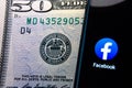 Facebook app on a screen next to 50 dollar bill. Conceptual: comparison of Facebook company app to Federal Reserve Systems emblem