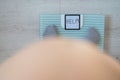 Faceless fat woman measures weight on a bathroom scale. Top view of a large naked female belly and feet in gray socks on