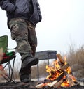 Man get warm by the fire in autumn or winter with first snow, authentic extreme camping in cold cloudy overcast weather
