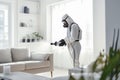 Faceless pest control worker in a protective suit sprays insect poison in a living room