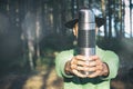 Faceless portrait of tourist hikerl man in felt hat holding stainless thermos before his face on woods background. Adventurer on