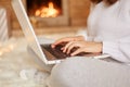 Faceless unknown woman sitting on floor on soft carpet at fireplace with laptop and working, typing, wearing white sweater and