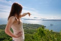 Faceless woman points hand at sea below, stands against panoramic view of green forest and ocean