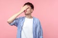 Facepalm. Portrait of distressed brown-haired man in casual striped shirt standing with hand on head. indoor studio shot, pink