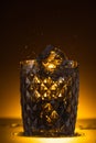 Faceted glass with ice cube and splashing vodka in dark with warm back light. Royalty Free Stock Photo