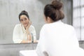 Facial Beauty.Beautiful smiling young Asian woman in bathrobe applying moisturizer cream on her pretty face and looking to mirror Royalty Free Stock Photo