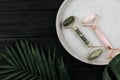 Facial roller from crystal rose quartz and massage tool jade Gua sha on the tray on black background with palm leaves.