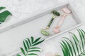 Facial roller from crystal rose quartz and massage tool jade Gua sha on the tray on grey background with palm leaves.