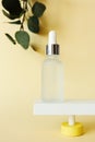 Facial serum bottle standing on abstract pedestal on pastel yellow background with copy space and defocused eucalyptus leaves