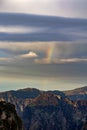 A faint rainbow arching above dramatic mountain peaks under a cloudy sky at sunset Royalty Free Stock Photo