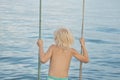 Fair-haired boy rides rope swing on sea background. Carelessness. Happy childhood