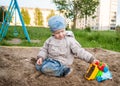 A fair-haired boy of three years in a blue hat and jeans, in a light jacket plays in the sandbox with toys