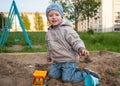 A fair-haired boy of three years in a blue hat and jeans, in a light jacket plays in the sandbox with toys Royalty Free Stock Photo