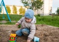 A fair-haired boy of three years in a blue hat and jeans, in a light jacket plays in the sandbox with toys Royalty Free Stock Photo
