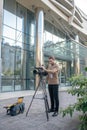 Fair-haired cameraman fixing camera to tripod in front of modern building