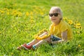 Fair-haired little child boy in yellow t-shirt sits on green grass lawn. Kid weaves wreath of dandelions sitting in field Royalty Free Stock Photo