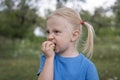 Fair-haired little girl in blue t-shirt eats green apple outside. Child walks in park or garden. Close-up portrait of little girl Royalty Free Stock Photo