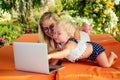 Fair-haired one year old female kid looking at a laptop cartoon with mother blonde freelancer surfer on sunbeds resort