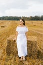 A fair-haired young woman in a white dress in a field with stacks of straw is enjoying a summer day Royalty Free Stock Photo