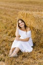 A fair-haired young woman in a white dress in a field with stacks of straw enjoys a summer day and smiles Royalty Free Stock Photo